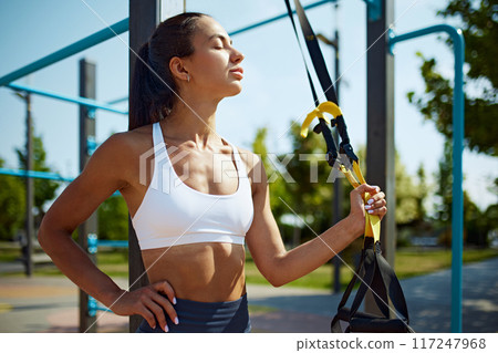 Standing with TRX strap in hand, fit woman taking breath deeply before intensive training session on sunny morning outdoor. 117247968