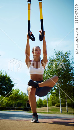 Motivation. Balance. Using suspension straps, woman intensifies her training by sitting in yoga pose, working on her core and stability outdoor. Motivation. Balance. Using suspension straps, woman intensifies her training by sitting in yoga pose, working on her core and stability outdoor. 117247969