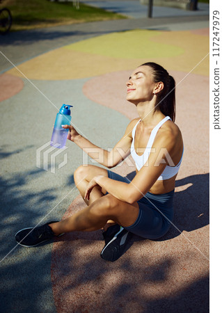 Young woman sitting outdoor and drink water in break between training sessions. Workout at outdoor gym on sunny morning for productivity. Young woman sitting outdoor and drink water in break between training sessions. Workout at outdoor gym on sunny morning for productivity. 117247979