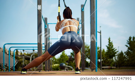 Engaged in high-intensity workout, woman doing stretching exercises for legs fully utilizing the TRX straps on sunny morning. Engaged in high-intensity workout, woman doing stretching exercises for legs fully utilizing the TRX straps on sunny morning. 117247991
