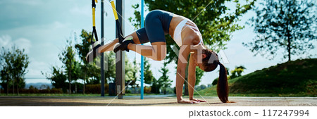 Using suspension straps, woman intensifies her training by lifting her legs, working on her core and stability on sunny day outdoor. Using suspension straps, woman intensifies her training by lifting her legs, working on her core and stability on sunny day outdoor. 117247994