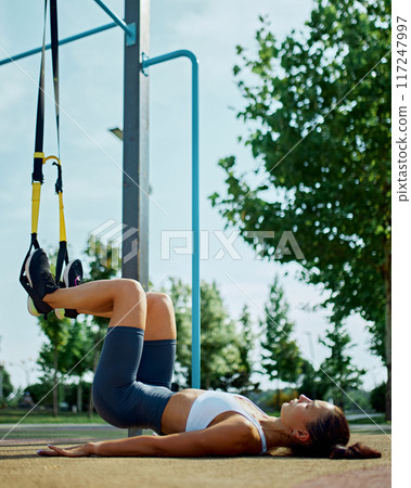 Young beautiful woman, doing exercises for legs, upper body using TRX loops on sunny summer day. Morning workout at park. Young beautiful woman, doing exercises for legs, upper body using TRX loops on sunny summer day. Morning workout at park. 117247997