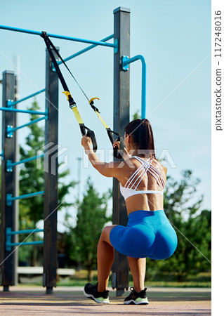 Full body of athlete woman workout outdoors with suspension system, doing stretching and strength exercises using special straps. Full body of athlete woman workout outdoors with suspension system, doing stretching and strength exercises using special straps. 117248016
