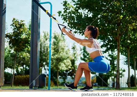 Full body of athlete woman workout outdoors with suspension system, doing stretching and strength exercises using special straps. Full body of athlete woman workout outdoors with suspension system, doing stretching and strength exercises using special straps. 117248018