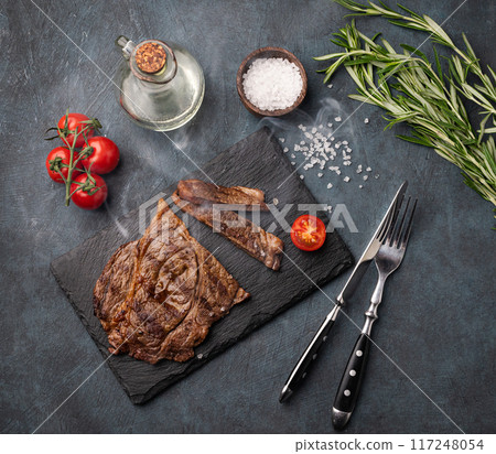 BBQ grilled beef steak with smoke, tomatoes, rosemary and spices on black slate on dark  background. 117248054