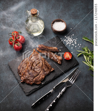 BBQ grilled beef steak with smoke, tomatoes, rosemary and spices on black slate on dark background. 117248055