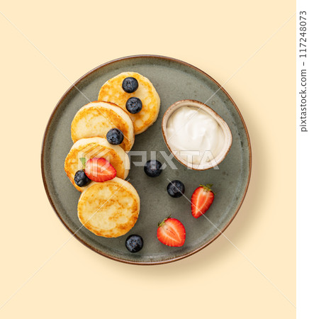 Cottage cheese pancakes with sour cream, blueberries and strawberries in a green plate  on a yellow background. Healthy and delicious breakfast. Top view 117248073