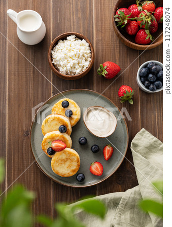 Cottage cheese pancakes with sour cream, milk, blueberries and strawberries on a dark wooden background. Healthy and delicious breakfast. Top view Cottage cheese pancakes with sour cream, milk, blueberries and strawberries on a dark wooden background. Healthy and delicious breakfast. Top view 117248074