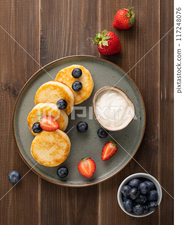 Cottage cheese pancakes with sour cream, blueberries and strawberries on a dark wooden background.  117248076