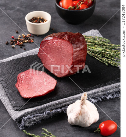 Seasoned smoked beef with  slices on black slate on dark background with fresh vegetables and herb. 117248094
