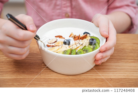 The hands of a girl in a pink shirt hold a bowl of yogurt, granola and fresh fruit on a wooden table The hands of a girl in a pink shirt hold a bowl of yogurt, granola and fresh fruit on a wooden table 117248408