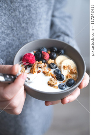 a young girl in a gray sweater is holding a bowl of vegetarian yogurt, granola and fresh blueberry  117248413