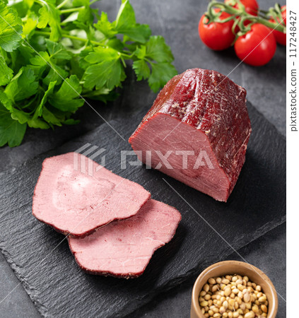Seasoned smoked beef with  slices on black slate on dark background with fresh vegetables and herb. 117248427