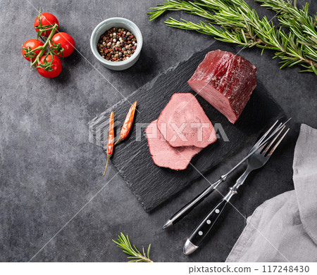 Juicy baked roast beef on a slate board with tomatoes, pepper and fresh herbs on a dark  background. 117248430