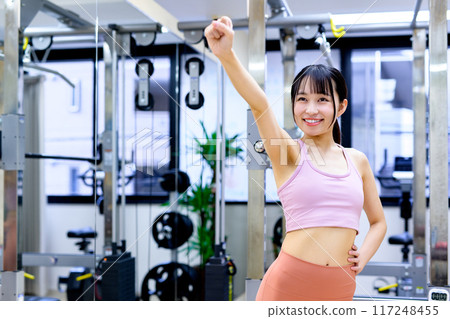Smiling young woman posing in a sports gym, diet, personal gym, muscle training, fist pump Smiling young woman posing in a sports gym, diet, personal gym, muscle training, fist pump 117248455