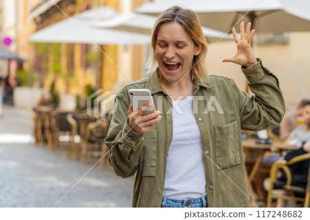 Happy young girl tourist using smartphone celebrating win good message news on urban city street 117248682