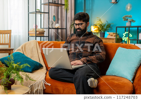 Happy young Indian man typing working on laptop browsing internet using apps sitting on home sofa 117248703