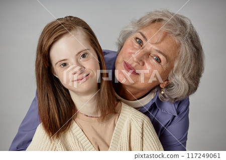 Portrait of young girl with Down syndrome and elderly woman smiling and embracing each other warmly against neutral background Portrait of young girl with Down syndrome and elderly woman smiling and embracing each other warmly against neutral background 117249061