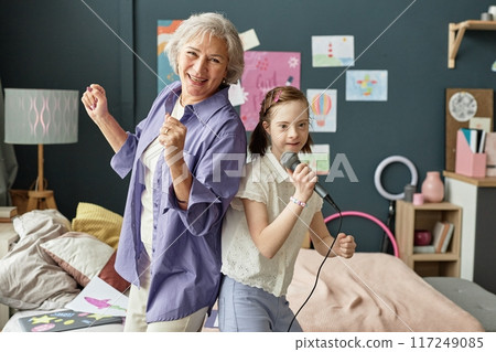 Elderly woman and young girl singing into microphone in cozy bedroom surrounded by drawings and artworks creating joyful atmosphere Elderly woman and young girl singing into microphone in cozy bedroom surrounded by drawings and artworks creating joyful atmosphere 117249085