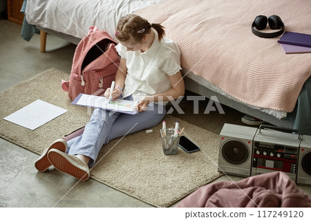 Young girl sitting on floor drawing artwork with pencil and paper placing near radio on rug next to bed and backpack 117249120