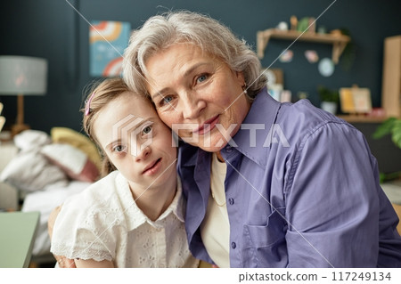 Portrait of senior woman with grey hair and young girl with Down syndrome sitting closely in cozy room Portrait of senior woman with grey hair and young girl with Down syndrome sitting closely in cozy room 117249134