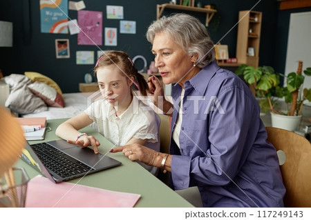 Senior woman assisting young girl with task on laptop in cozy room surrounded by various decorations and plants Senior woman assisting young girl with task on laptop in cozy room surrounded by various decorations and plants 117249143