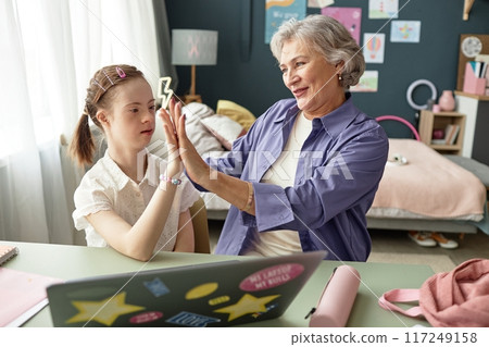 Elderly woman and young girl engaging in high-five in cozy bedroom with personal items. Laptop on desk symbolizes modern connection and interaction Elderly woman and young girl engaging in high-five in cozy bedroom with personal items. Laptop on desk symbolizes modern connection and interaction 117249158