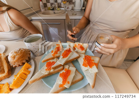 A woman is cutting bread and putting jelly on it. The table is set with a variety of food items, including sandwiches, fruit, and a jar of jelly. The woman is wearing a dress. A woman is cutting bread and putting jelly on it. The table is set with a variety of food items, including sandwiches, fruit, and a jar of jelly. The woman is wearing a dress. 117249734