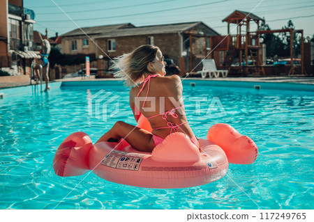 A woman is sitting on a pink flamingo float in a pool. The scene is bright and cheerful, with the woman enjoying her time in the water. 117249765