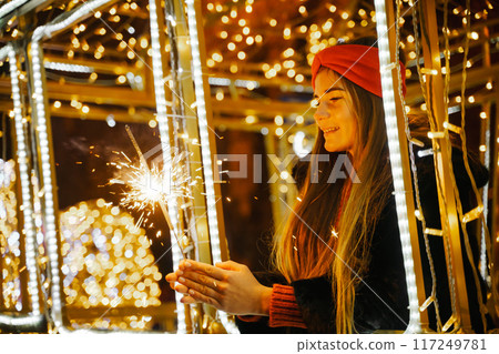 Woman holding sparkler night while celebrating Christmas outside. Dressed in a fur coat and a red headband. Blurred christmas decorations in the background. Selective focus Woman holding sparkler night while celebrating Christmas outside. Dressed in a fur coat and a red headband. Blurred christmas decorations in the background. Selective focus 117249781