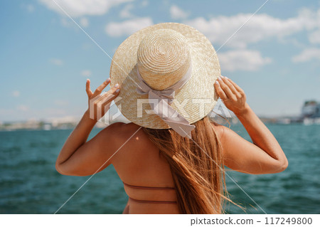 A woman in a swimsuit sits with her back holding a hat, looks at the ocean, sunny day, relaxes. A woman in a swimsuit sits with her back holding a hat, looks at the ocean, sunny day, relaxes. 117249800