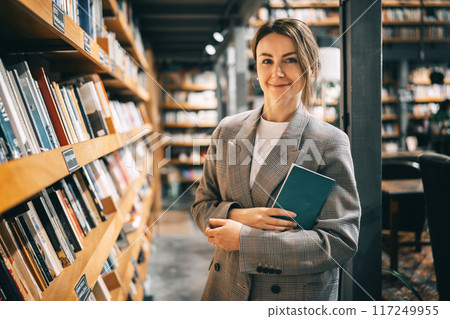 Young smiling woman stands amidst towering bookshelves in a cozy library 117249955