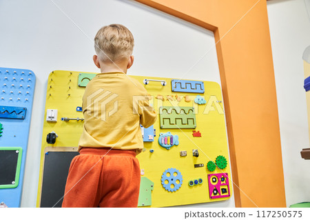 A child is playing a wooden maze on the wall. Educational logic toys for kids. An unrecognizable child with his back to the camera with a copy space. High quality photo A child is playing a wooden maze on the wall. Educational logic toys for kids. An unrecognizable child with his back to the camera with a copy space. High quality photo 117250575