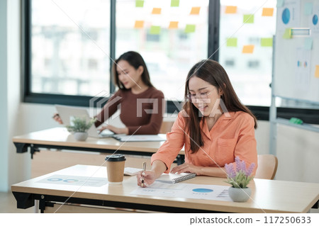 Two women are sitting at desks, one of them writing in a notebook 117250633