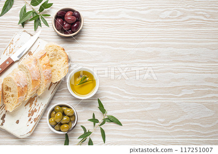 Sliced fresh ciabatta on cutting kitchen board, green and brown olives, olive oil with rosemary, olive tree branches on white wooden rustic background top view 117251007