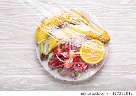 Assorted different fruit and berries on white plate wrapped in plastic clear film top view on white wooden rustic table background 117251012