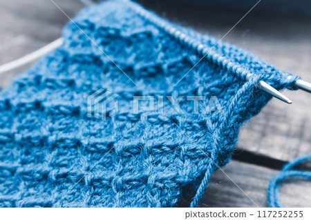 Blue wool yarn pattern with knitting needles. Blue knitting wool on grunge wooden background. Woven crochet close up. Knitting hobby. Knitwear concept. Needlework background.  117252255