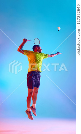 Full-length image of young concentrated badminton player, man hitting shuttlecock in a jump against gradient blue pink background in neon light 117252649