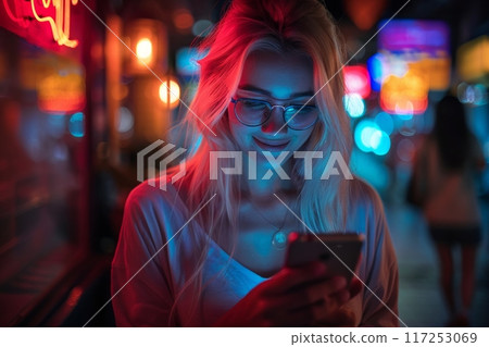 young woman with blonde hair and glasses smiles as she looks at her phone, illuminated by the vibrant neon lights of the city at night. 117253069