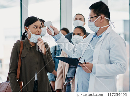 Travel medical healthcare worker testing covid temperature at airport using infrared thermometer. Professional doctor doing a coronavirus check up on a woman at an office entrance Travel medical healthcare worker testing covid temperature at airport using infrared thermometer. Professional doctor doing a coronavirus check up on a woman at an office entrance 117254493