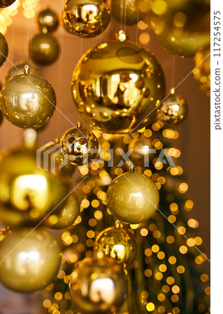 Many large golden Christmas balls look hanging against the background of the Christmas interior. Festive background in golden awards, bokeh, selective focus Many large golden Christmas balls look hanging against the background of the Christmas interior. Festive background in golden awards, bokeh, selective focus 117254575