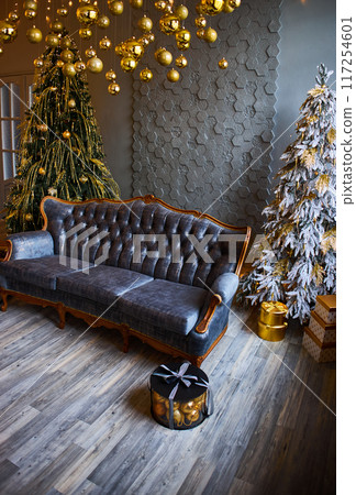 Luxurious interior of the room for Christmas and New Year. Living room in gray and gold tones with a sofa, many gifts, golden balls, ice lights, two Christmas trees Luxurious interior of the room for Christmas and New Year. Living room in gray and gold tones with a sofa, many gifts, golden balls, ice lights, two Christmas trees 117254601
