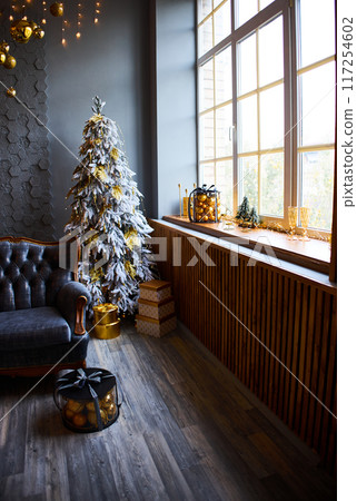 Luxurious interior of the room for Christmas and New Year. Living room in gray and gold tones with a sofa, many gifts, golden balls, ice lights, two Christmas trees Luxurious interior of the room for Christmas and New Year. Living room in gray and gold tones with a sofa, many gifts, golden balls, ice lights, two Christmas trees 117254602