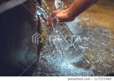 A man collects crystal clear mineral water from a spring into a transparent glass bottle. The hand brings the bottle to a stream of clear, clean water to fill it. Water with bubbles in bottles. 117255460