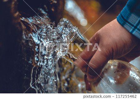 A hand brings a transparent bottle to a spring of drinking water. A man collects mineral water from a spring in the autumn forest. The traveler stopped at a spring to drink and collect clean water. A hand brings a transparent bottle to a spring of drinking water. A man collects mineral water from a spring in the autumn forest. The traveler stopped at a spring to drink and collect clean water. 117255462