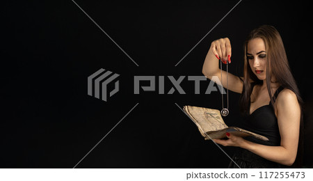 Halloween divination. Copyspace. A black witch tells fortunes over a book with a magic talisman on a chain in her hand. In the semi-darkness on Halloween, the witch will perform her dark ritual. 117255473