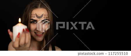 On Halloween, the witch, smiling sweetly, holds out a burning candle to the camera. Copy space. On a black background, a woman in the form of a witch with magical symbols on her face holds a candle. 117255510
