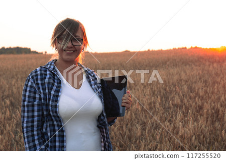 Cute smiling farmer woman with a tablet in her hands looks at the camera on the background of a wheat field at sunset. A beautiful woman smiles in the rays of the setting sun in a wheat field. Cute smiling farmer woman with a tablet in her hands looks at the camera on the background of a wheat field at sunset. A beautiful woman smiles in the rays of the setting sun in a wheat field. 117255520