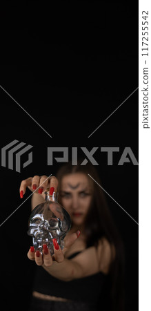 vertically. On outstretched arms on Halloween night, a witch holds a glass skull in the dark. Copyspace. Studio shot. Portrait. A woman holds out a glass skull from a twilight Halloween evening. 117255542