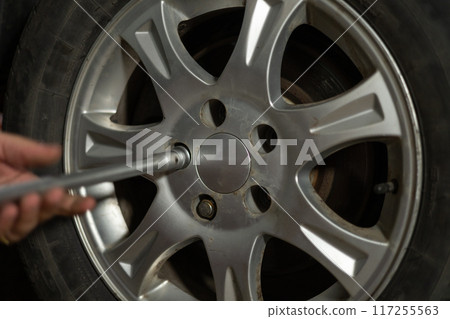 A man in a garage unscrews a car wheel with a key. The car stands on a jack while a man repairs a wheel. Replacing a flat tire on a car in the garage. The concept of do-it-yourself transport repair. A man in a garage unscrews a car wheel with a key. The car stands on a jack while a man repairs a wheel. Replacing a flat tire on a car in the garage. The concept of do-it-yourself transport repair. 117255563